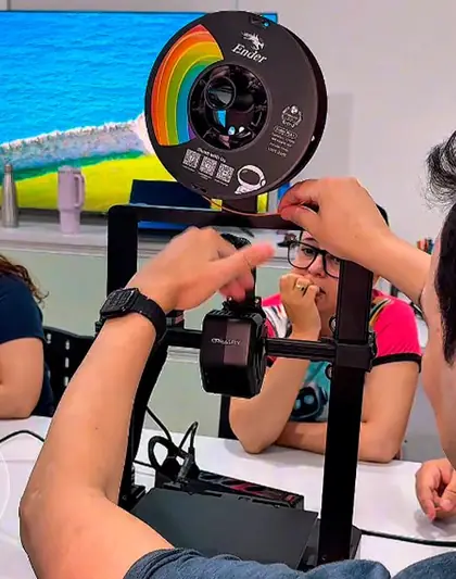 Mãos ajustam o filamento de uma impressora 3D enquanto uma participante observa